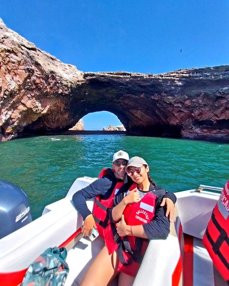 A smiling couple in life jackets on a boat with a natural rock arch in the background during the Paracas Full Day tour