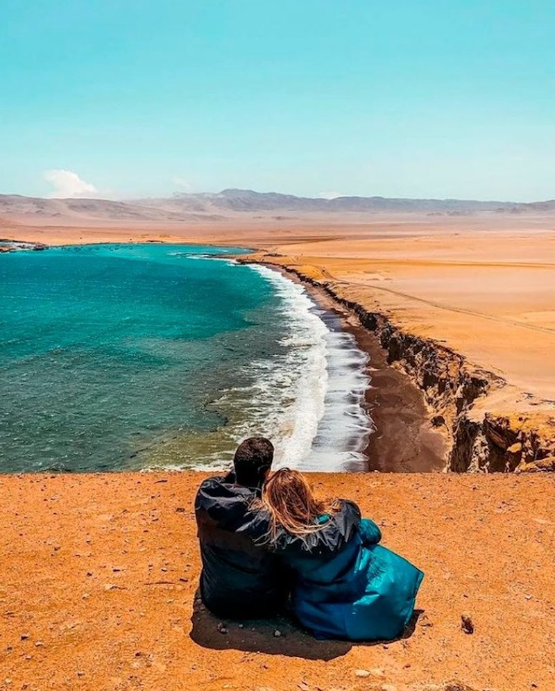 A couple sitting on a sandy cliff overlooking the waves and a beautiful beach in the Paracas National Reserve, Paracas Full Day tour.