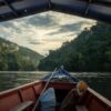 A boat navigating through a river flanked by dense forest under a cloudy sky in Manu National Park