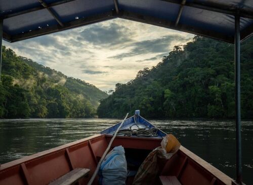 A boat navigating through a river flanked by dense forest under a cloudy sky in Manu National Park